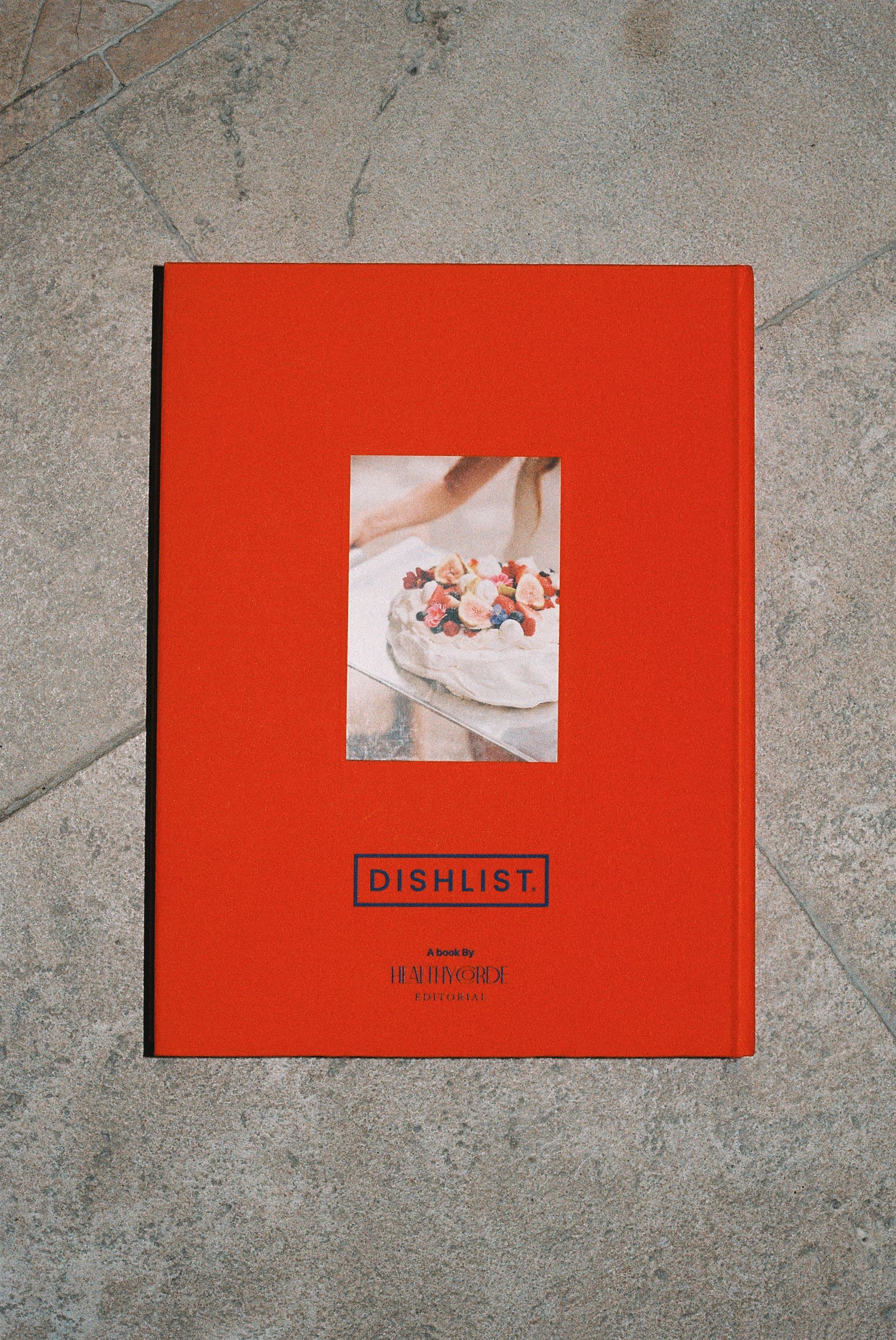 Contracubierta del libro Dish List vista desde arriba sobre piedra: foto inset de una pavlova con frutos rojos, el wordmark DISHLIST y la firma de Healthycorde Editorial.